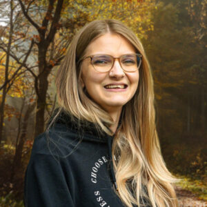 Lena ist in einer Portrait Ansicht zu sehen, sie lächelt in die Kamera. Der Hintergrund ist ein Wald in herbstlichen Farben.